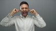 © Krakenimages.com - Hispanic man wearing white shirt plugs ears with fingers in front of grey background, expressing frustration and blocking sound.