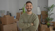 © Krakenimages.com - Young hispanic man smiling indoors with arms crossed standing among cardboard boxes and plants suggesting moving into a new home with a cozy atmosphere.