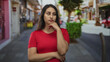 © Krakenimages.com - Woman with hand on chin and crossed arms standing on a street in a city near shops, wearing red top, looking sideways; thoughtful reflection decision.