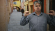 © Krakenimages.com - Middle age man points thumbs to himself with clenched mouth and visible hands in narrow street, archway behind; annoyance resignation.