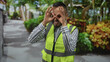 © Krakenimages.com - Man making hand binoculars gesture with bare hands and high visibility vest on street with park foliage; playful curiosity.