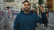 © Krakenimages.com - Man showing three fingers with raised hand, beard visible and blue hoodie, standing on busy city street by traffic lights and buildings; confidence casual.