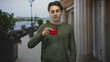 © Krakenimages.com - Man holding red mug by its handle with right hand, wearing green long sleeve shirt, standing before tall building columns; quiet contemplation.