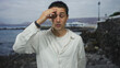 © Krakenimages.com - Young hispanic man in white shirt stands thoughtful on a beach with a rocky seaside backdrop under a cloudy sky, conveying a sense of outdoor contemplation and tranquility.