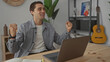 © Krakenimages.com - Young man in casual attire using laptop at home in cozy living room with guitar and bookshelves in background, engaging in a virtual meeting with a focused expression.