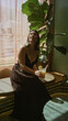 © Krakenimages.com - Woman brunette holding glass with lemon garnish seated at table in building interior by window and large plant; calm leisure.