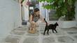 © Krakenimages.com - Young hispanic brunette woman crouching on a sunlit mykonos street reaches hand to pet a black cat while smiling; serenity and connection.