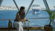 © Krakenimages.com - Woman brunette holding cocktail glass by window, sitting on bench in building with cruise ship view, striped top and white pants, straw bag beside her; serenity.