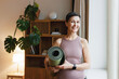 © shurkin_son - Happy energized female of 50s standing in her living-room in activewear and holding rolled mat, enthusiastic about her morning pilates training at home and its results as her toned muscles