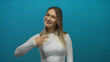 © Krakenimages.com - Woman pointing at herself with a thumbs-up gesture against a blue wall, showing confidence and positivity in a youthful, casual manner, isolated in a bright studio setting.