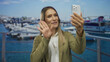 © Krakenimages.com - Woman smiling while holding smartphone during video call on a port, showcasing outdoor seaside setting with boats and clear blue sky in the background.