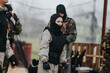 © qunica.com - A woman in camouflage gear and protective vest prepares for a paintball match. Outdoor field with teammates and netting provides the backdrop.