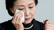 © bephoto - 灰色背景で涙を拭う東アジアの女60代、喪服と数珠の静かな悲しみ写真