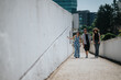 © qunica.com - Three friends walk along a sunlit white wall on an urban pathway. They chat and stroll together in a casual city setting.