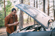 © Anna - A frustrated man with a phone in his hands near a broken car with an open hood far outside the city in the woods.