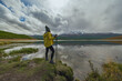 © ANDREY - Back view of female hiker in yellow raincoat and beanie with trekking poles standing on shore of scenic mountain lake with snow capped peaks reflection under dramatic cloudy autumn sky.