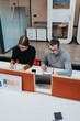 © qunica.com - Two colleagues sit at a white desk, writing in notebooks in a bright office. A contemporary workspace with dividers, phones, and office furniture behind them.