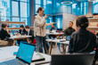 © qunica.com - A woman speaks into a microphone in a bright coworking space while colleagues listen. The scene shows a collaborative work environment with laptops and casual seating.