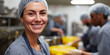© Dinali - Smiling white woman in hairnet and apron working in a food production facility, with colleagues in the background Concept of food industry, teamwork, and dedication
