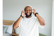 © Wavebreak Media - Man smiling and holding headphones while listening with smartphone in bright bedroom wearing beanie