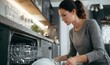 © arayabandit - A focused young woman loads clean white dishes and glasses into a modern, built-in dishwasher, symbolizing household chore and kitchen organization.