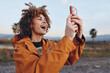 © SHOTPRIME STUDIO - A happy woman with curly hair wearing an orange jacket smiles broadly while holding a smartphone outdoors during sunset. The woman enjoys capturing a moment in nature.