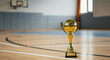 © Artyom - Golden basketball trophy standing on a hardwood court near a hoop, symbolizing championship victory in an indoor gymnasium