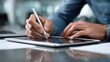 © Zabi - A man's hands delicately hold a silver stylus pen, writing on a tablet screen with a laptop in the blurred background.