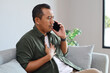 © ArvStd - An adult man is talking on his cell phone while sitting on the sofa at home with a shocked expression while holding his chest.