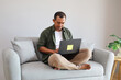 © ArvStd - A handsome man relaxes comfortably at home, sitting cross-legged on the sofa, focusing on his laptop with a relaxed smile, working from home.