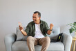 © ArvStd - A man sitting on a sofa happily celebrating success at home with a laptop and notebook, showing enthusiasm and productivity in a comfortable environment, enjoying a dynamic and modern lifestyle.