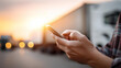 © Denis Yevtekhov - Logistics professional overseeing truck fleet and delivery on smartphone app, loading dock operations with sunset sky, transportation management technology, defocused vehicle backg