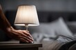 © whitestorm - Close up of a hand reaching to turn on a bedside lamp in a dark bedroom, light illuminating the textured lampshade and creating a warm, inviting atmosphere.