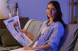 © Pixel-Shot - Young woman reading newspaper on sofa at home in evening