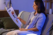 © Pixel-Shot - Young woman reading newspaper on sofa at home in evening