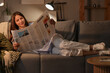 © Pixel-Shot - Young woman reading newspaper on sofa at home in evening