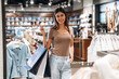 © standret - Young woman with long dark hair smiles while holding shopping bags inside a clothing store, racks of apparel and accessories visible in the background