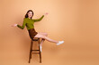 © deagreez - Young woman sits on a wooden stool in a beige studio smiling with a green top brown shorts and white sneakers creating a casual fashion lifestyle stock image