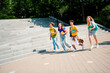 © deagreez - Group of cheerful young students walking outdoors on a sunny day holding book and backpacks near urban stairway