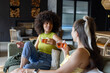 © Wavebreak Media - Diverse female friends wearing lime yellow tops, chatting on cozy sofa holding glass mugs at home