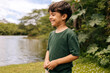 © kleberpicui - Brazilian boy smiling near a calm lake surrounded by greenery, portraying joyful childhood, outdoor freedom and connection with nature in a peaceful environment.