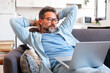 © simona - Relaxed mature man stretching arms while working and watching screen laptop sitting comfy on the sofa at home in technology leisure online activity or business time