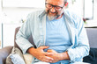 © simona - Sick bearded man wearing glasses suffering from stomach ache, holding his belly and feeling discomfort. Upset man with abdominal pain, gastritis symptom, indigestion problem, isolated on gray studio