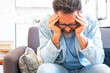 © simona - Sad and frustrated 50s man suffering from stress, depression at home, sitting on sofa, touching head, feeling headache, migraine, dealing with loss, grief, trauma. Nervous breakdown, crisis concept
