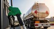 © Desingora - Closeup of green fuel nozzle at gas station with large tanker truck in background under bright sunlight