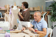 © JackF - Experienced tailor sews clothes against the background of his assistant, who checks a sample of the dress on a mannequin. Male tailors work on ordering clothes in the workshop