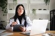 © Studio Marmellata - A woman in a white lab coat and glasses works on a tablet with a stylus, with a laptop open on her desk.