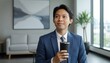 © MopoWe - Young thoughtful asian businessman in a sophisticated blue suit holds a black insulated cup looking out of an office window