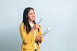 © Ryan - Smiling Indonesian woman wearing yellow blazer and white shirt, holding green clipboard and raising pen while looking up at copy space, isolated on white background.