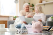 © Pixel-Shot - Piggy bank with refund form and coins on table of senior couple at home, closeup